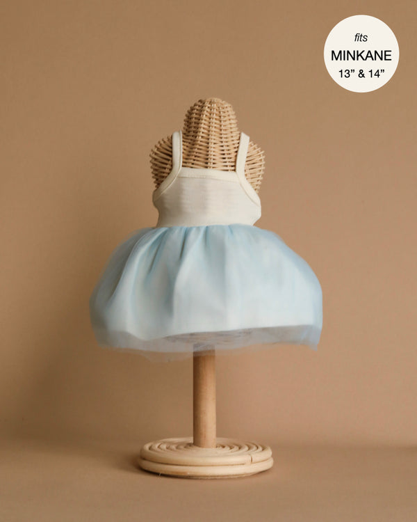 A light-colored woven mannequin displays the Rosella Tutu in Sky Blue, a sleeveless dress featuring a white bodice and a light blue tulle skirt. Designed to fit 13- and 14-inch Minikane dolls, as mentioned in the upper right corner, this charming outfit is hand washable. The background is a plain, light brown.