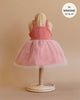 A child's dress displayed on a wicker dress form, perfect for Minikane dolls. The Rosella Tutu in Marsala features a sleeveless pink bodice and a voluminous soft pink tulle skirt. Text in the top right corner reads, "fits 13” & 14”." The background is a plain, neutral color, showcasing its designed-in-France elegance.