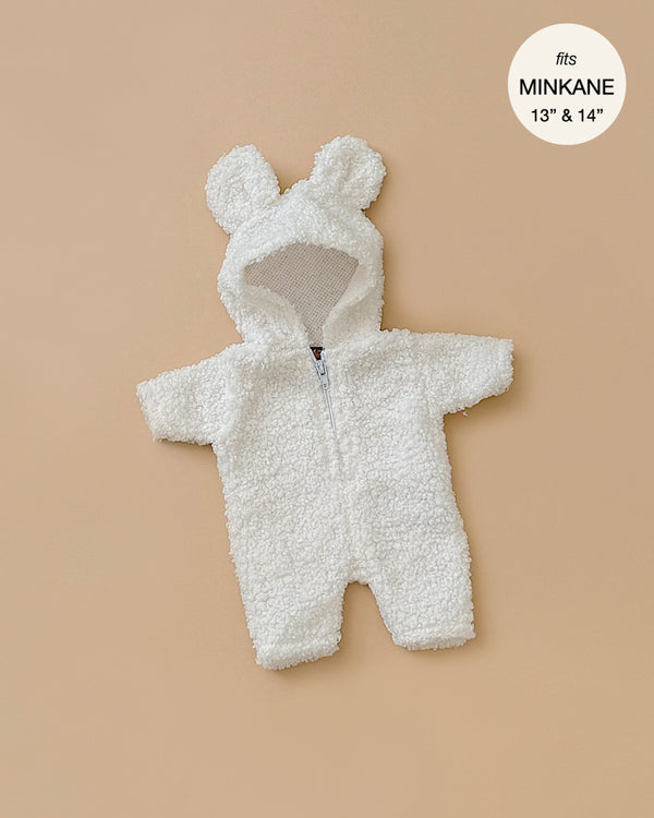 A cozy and adorable Winnie Jumpsuit in Ecru Bouclette, designed as a fluffy white onesie with a hood featuring bear ears, is elegantly laid flat on a beige background. Perfectly suited for 13" and 14" Minikane dolls, this onesie embodies charming details and proudly states "fits Minikane," reflecting its made-in-France craftsmanship.