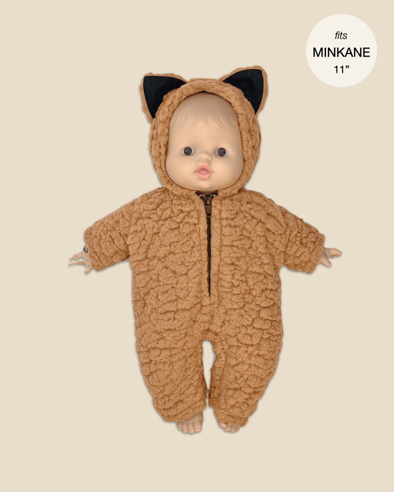 A doll dressed in a snug Brown Sugar Sherpa Halloween Onesie, mimicking the style of your favorite sherpa jumpsuit, showcases an adorable hood with small black cat ears. Designed for 11-inch Minikane Babies, it stands out against a neutral backdrop.