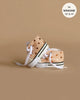 A pair of miniature peach-colored Converse style sneakers with white laces and white soles, adorned with gold sequins. The background is a neutral beige color. A circular label in the top right indicates the shoes fit Minikane Gordis dolls sizes 13 inches and 14 inches, and mentions "MINIKANE".