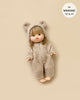 A doll dressed in the Minikane Doll Clothing | Winnie Beige Terry Jumpsuit, complete with a hood featuring bear ears, is laying on a neutral background. The Minikane doll has short brown hair and a peaceful expression. A text bubble reads, "fits MINIKANE 13" & 14". Made in France.