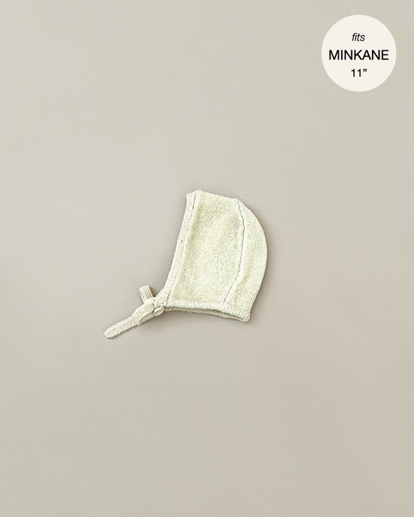 A small, light-colored baby bonnet with a tie string is displayed against a neutral background. A circular label in the top right corner reads "Minikane Doll Clothing | Élie Cream Knit Bonnet fits Minikane Babies 11''".