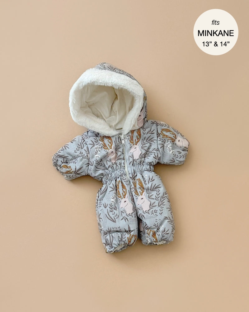 The Gigi Elaphe Ski Suit is a cozy baby snowsuit featuring a woodland animal print on a light background, complete with a hood, ideal for Minikane dolls. Made in France, the image includes text within a circle that reads "fits MINKANE 13'' & 14''." The suit is displayed on a beige background.