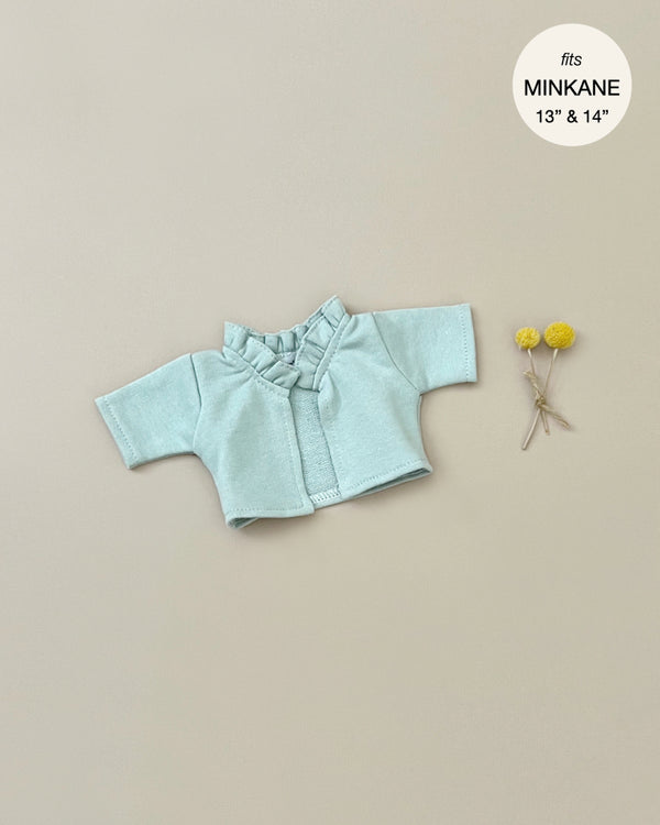 A Minikane Doll Clothing | Claudia Cardigan in Green Tea Fleece is displayed on a beige background. Two small yellow flowers are placed to the right of the cardigan. The text in the corner indicates that the cardigan fits 13" and 14" MINKANE dolls.