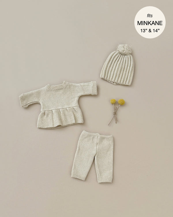 The Minikane Doll Clothing | Lina Set in Cream Knit includes a cream-colored knit outfit for Minikane Gordis dolls, featuring a peplum sweater, pants, and a ribbed hat with a pom-pom. Two small yellow flowers lie next to the Lina knitting set. A circular text label reads, "fits MINKANE 13" & 14"." The background is beige.