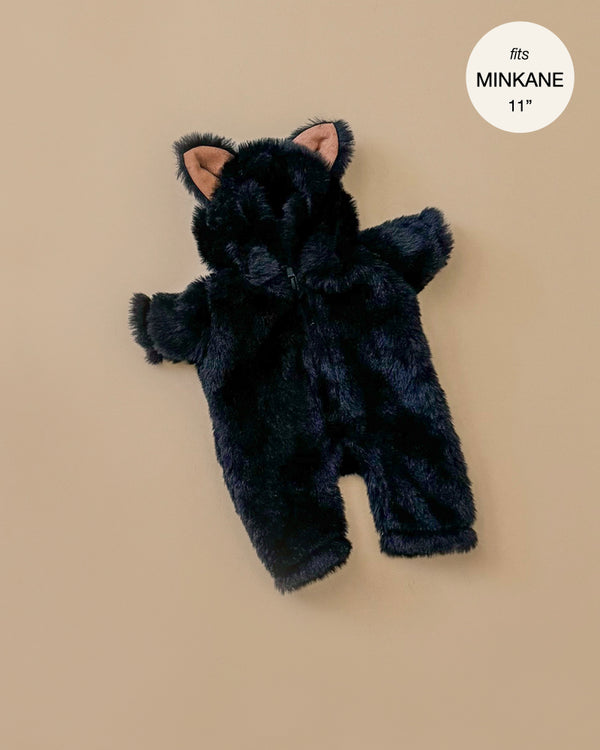 A small, black faux fur costume designed for an 11-inch Minikane doll is laid out on a beige background. The jumpsuit includes adorable cat ears on the hood and looks soft and fluffy. A circular label in the top right corner reads "fits MINKANE 11", making it ideal for the Halloween capsule collection.