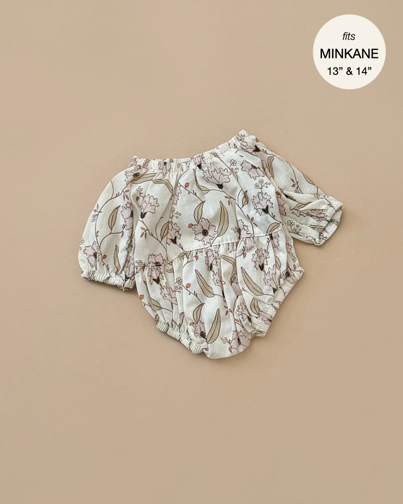 The Janelle Cotton Bertille Romper, designed for Minikane Gordis dolls sized 13" and 14", features a nature-inspired floral and leaf pattern, displayed on a beige background. A tag in the top right corner indicates it "fits MINKANE 13" & 14"." Made in France, this romper is hand washable for convenient care.