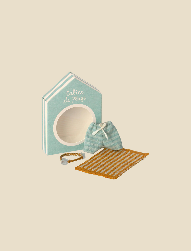 A small, house-shaped box labeled "Cabine de Plage" with an open circular window. In front of the box are a striped drawstring pouch, a striped mat, and a small seashell bracelet—all part of the Maileg Beach Set for Big Brother. The items are arranged on a beige background, creating your perfect vacation set.