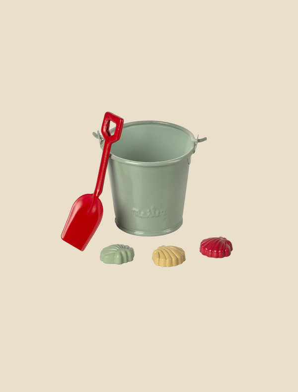 The Maileg Sand Toys set includes a light green metal bucket with a handle, paired with a red plastic shovel, creating an indispensable beach accessory for your vacation. In front of the bucket are three small, vibrant sand toys shaped like seashells in light green, yellow, and red hues. The scene is set against a plain beige background.