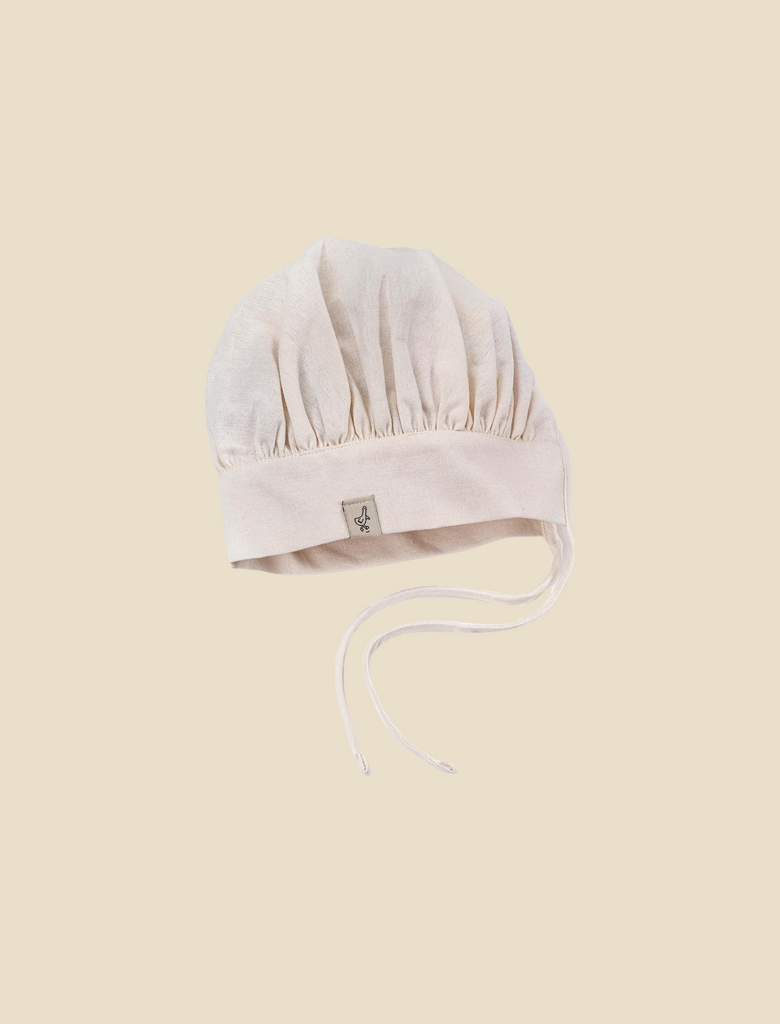 A soft, light beige baby Milton & Goose Chef's Hat with delicate stitching and fabric ties, displayed centered on a pale yellow background. The bonnet features a small tag with a logo on the side and resembles a handmade