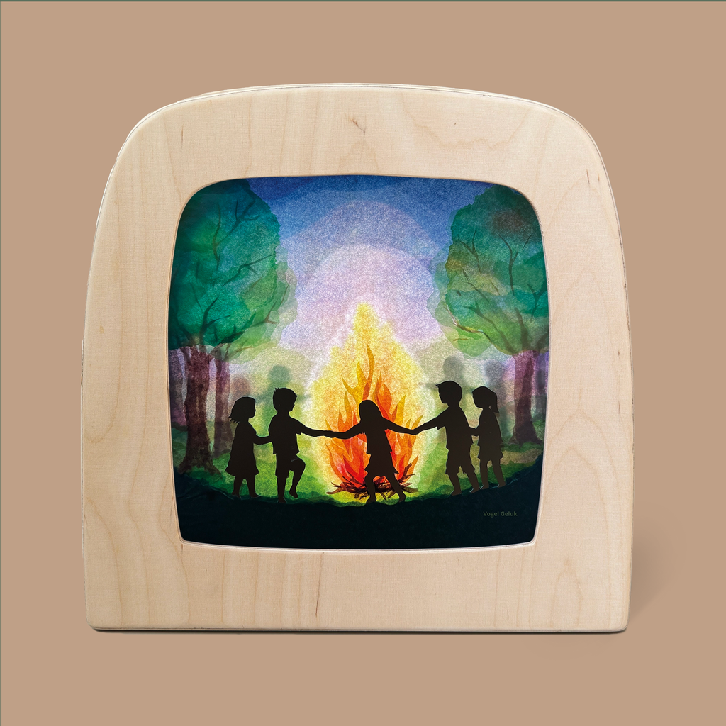 A St. Jans Fire Silhouette for the Toverlux Lamp features a wooden frame that displays an illustration of five Magic Silhouettes holding hands in a circle around a campfire, encircled by trees under a vibrant, starry night sky, all glowing as if crafted on translucent material.