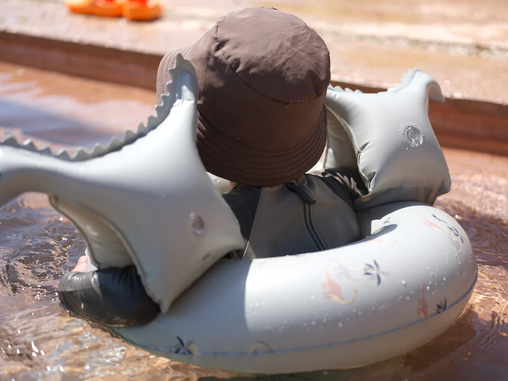 A small child, presumably 3-6 years old, wearing a brown wide-brim hat and a grey shark-themed flotation device with Inflatable Water Wings - Dinosaur Shape is sitting in shallow water on a sunny day. The child is partially submerged and facing away from the camera. The clear water reveals some steps in the background.