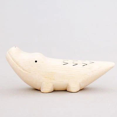 A simple wooden alligator toy crafted from Albizia wood, branded as "Handmade Tiny Wooden Safari Animals - Alligator." This light beige alligator features minimalistic details, including hand-painted black eyes and a few black zigzag lines on its back to suggest scales. The background is a plain gray.