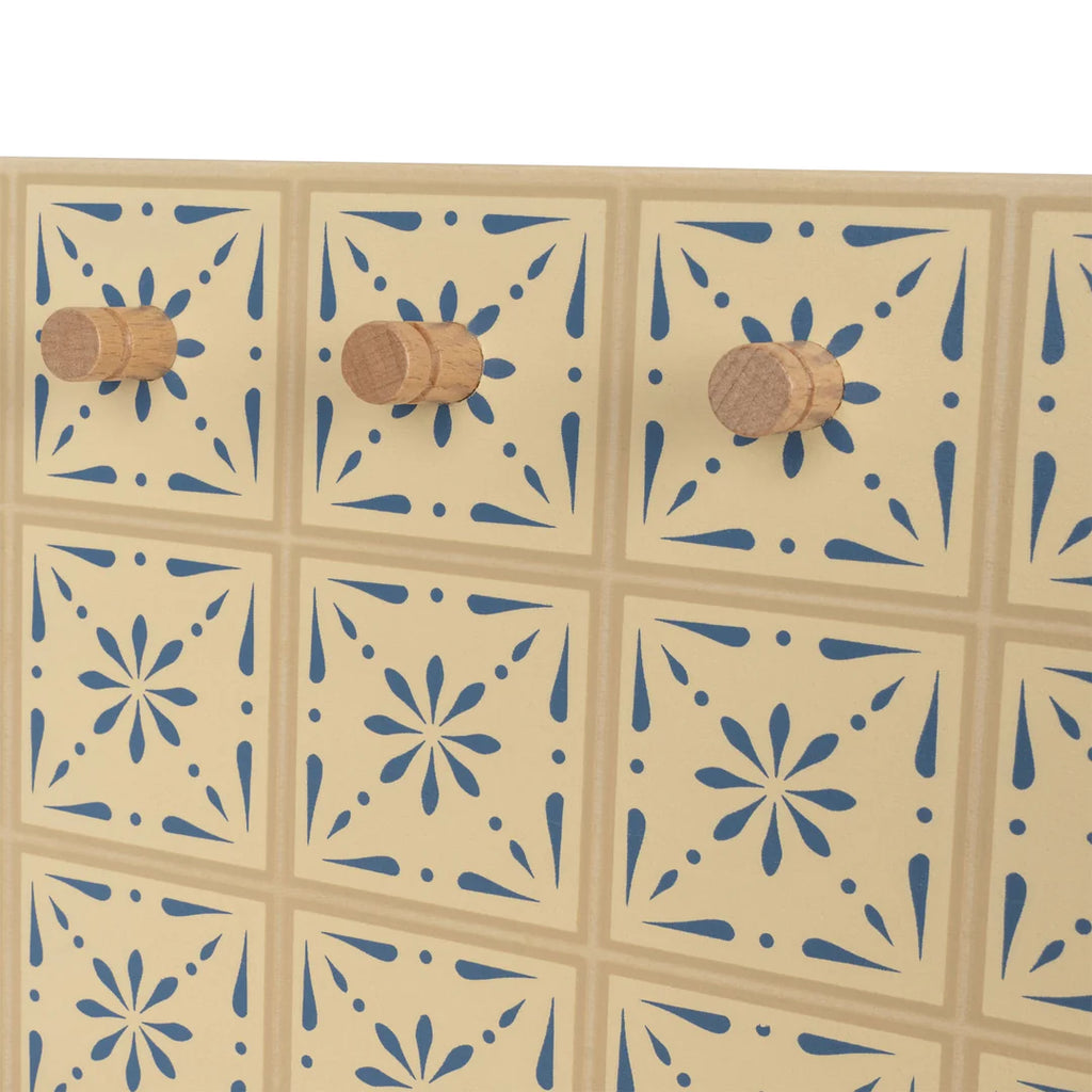 A wooden stove with a pattern of tan and blue tiles arranged in rows, each featuring a floral-like design, rests on a wooden table in the kitchen. Three wooden pegs are evenly spaced along the middle row of the stove.