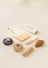 Introducing the Wooden Bakery Set: a delightful collection of wooden kitchen toys and play dough items, perfect for a children's game that promotes healthy eating habits. This set includes a cutting board, knife, various types of bread like croissant and donut, and a mysterious black object with an X mark. A rolled-up cloth with text ties it all together on a light-colored surface.