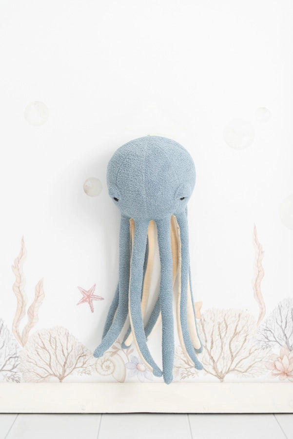 A soft blue Octopus Stuffed Animal hangs on a white Deco Wall decorated with subtle undersea illustrations featuring coral and bubbles, ideal for a children's room decoration.