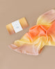 A vibrant, flowing silk scarf in shades of yellow, orange, and pink is laid out on a beige surface. Next to it, a folded version wrapped with a minimalist band labeled "PLAYSILK" showcases Sarah's Silk Enchanted Playsilk - Desert, emphasizing its role in imaginative play and its creation from natural materials.