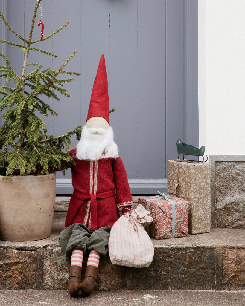 A Maileg Large Santa, with its long white beard and red outfit, sits on a doorstep next to a potted evergreen tree. It clutches a striped sack and sits beside two wrapped gifts, one topped with a small green sled—a charming decoration to bring holiday cheer.