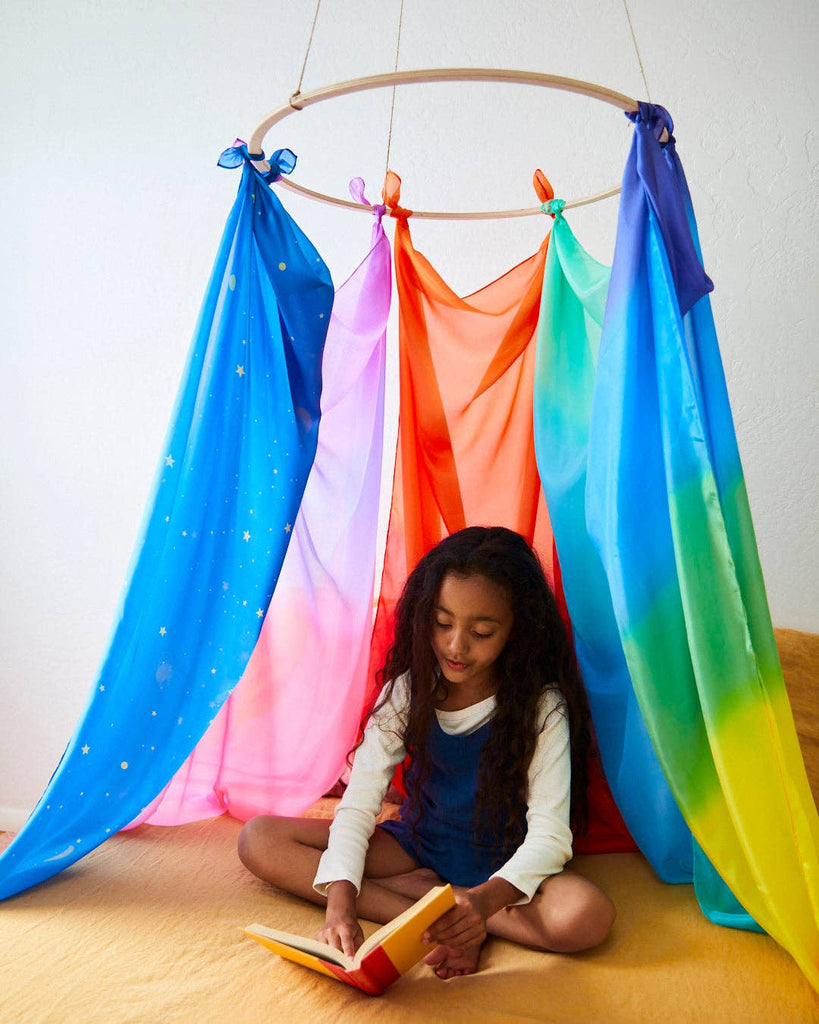 A young girl sits cross-legged on a bed, immersed in imaginative play as she reads a book. She is under a colorful canopy crafted from Sarah's Silk Giant Rainbow Playsilk, flowing gracefully from a circular frame above. The room exudes a calm and cozy atmosphere.