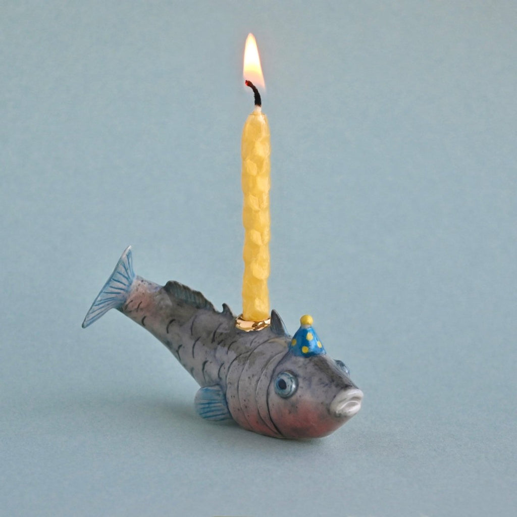 A whimsical hand-painted Fish Cake Topper shaped like a fish with a lit wick on its back, against a plain light blue background. The fish is painted with realistic details and wears a small party hat.