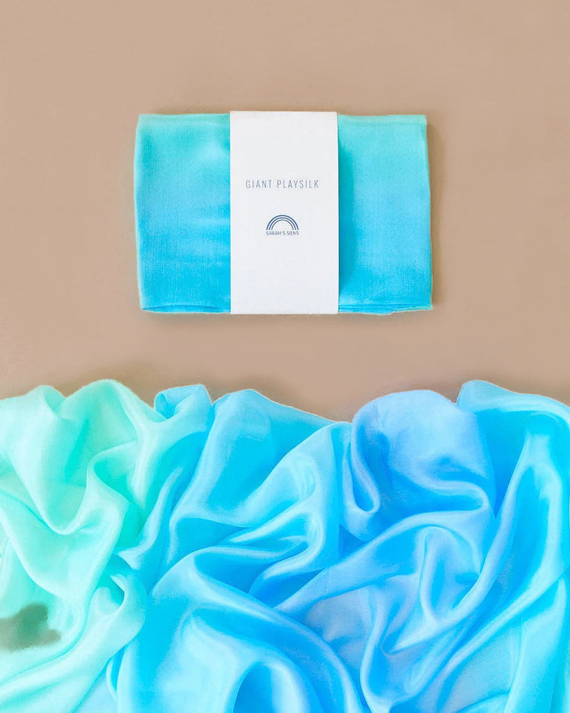 A flat lay of a folded, gradient blue and green silk fabric labeled "Sarah's Silk Giant Sea Playsilk" featuring a rainbow logo on a beige background. Below, the fabric is unfolded, encouraging imaginative play with its soft texture and vibrant colors.