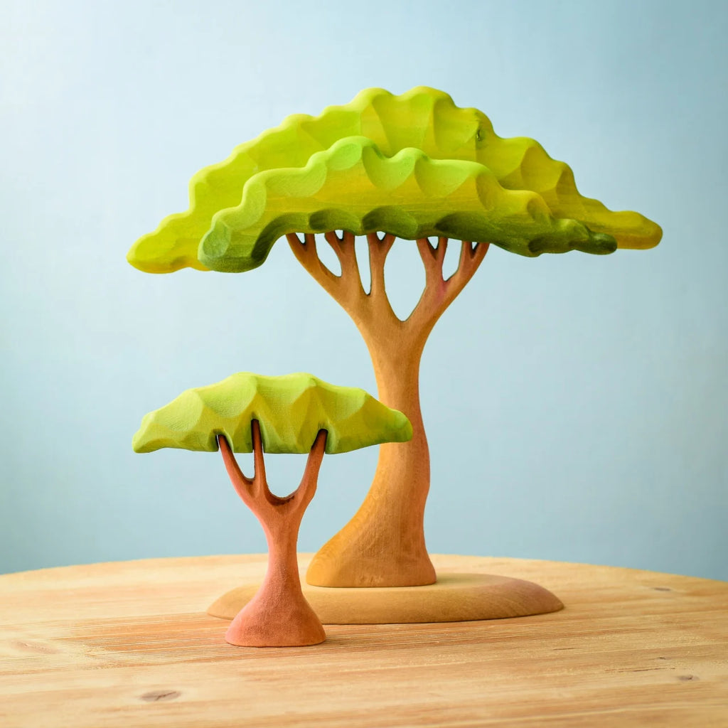 Two wooden toy trees from the Bumbu Wooden Acacia Tree Set sit on a light wooden surface against a soft blue background. The larger tree, inspired by Montessori and Waldorf designs, has a broad, wavy green canopy and a wide brown trunk. The smaller tree shares this eco-friendly aesthetic but features a thinner trunk and petite canopy.