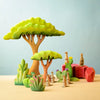 The Bumbu Wooden Acacia Tree Set is a vibrant diorama that showcases two large trees, various green plants, pink rock formations, and several small meerkat figurines on a beige surface against a light blue background. This handmade set exemplifies Montessori and Waldorf principles, offering eco-friendly toys that inspire imaginative play.