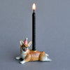 A whimsical hand-painted porcelain Corgi Cake Topper featuring a figurine of a smiling corgi wearing a party hat, supporting a lit black taper candle on a gray background.