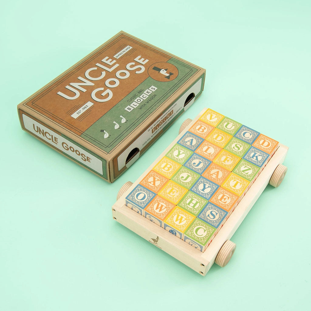 A set of Uncle Goose Classic ABC Blocks with Wagon is displayed. The packaging is on the left with "Uncle Goose" written in bold letters. The ABC blocks are arranged on a basswood wagon with wheels on the right, showing letters and numbers in various colors and patterns.