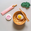 A wooden bowl with toy noodles, a toy egg, and chopsticks. Next to the bowl, there is a green leaf-shaped dish, a pink and white spiral fish cake toy for sensory play food, and a pink package labeled "kiko+ x oekgarol" containing chopsticks. Crafted from natural non-toxic materials, this Ramen Bowl Playset (ships in approximately one week) is perfect for imaginative fun.