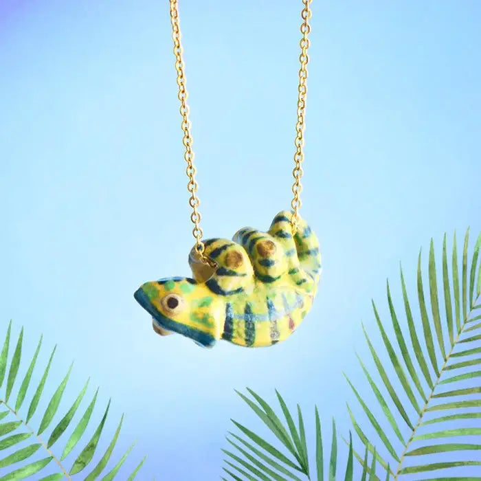 A whimsical Chameleon Necklace featuring a vibrant, green and yellow striped pendant hangs on a delicate 24k gold plated steel chain. The background is a soft blue gradient with green palm fronds accentuating the bottom corners, giving a tropical vibe.