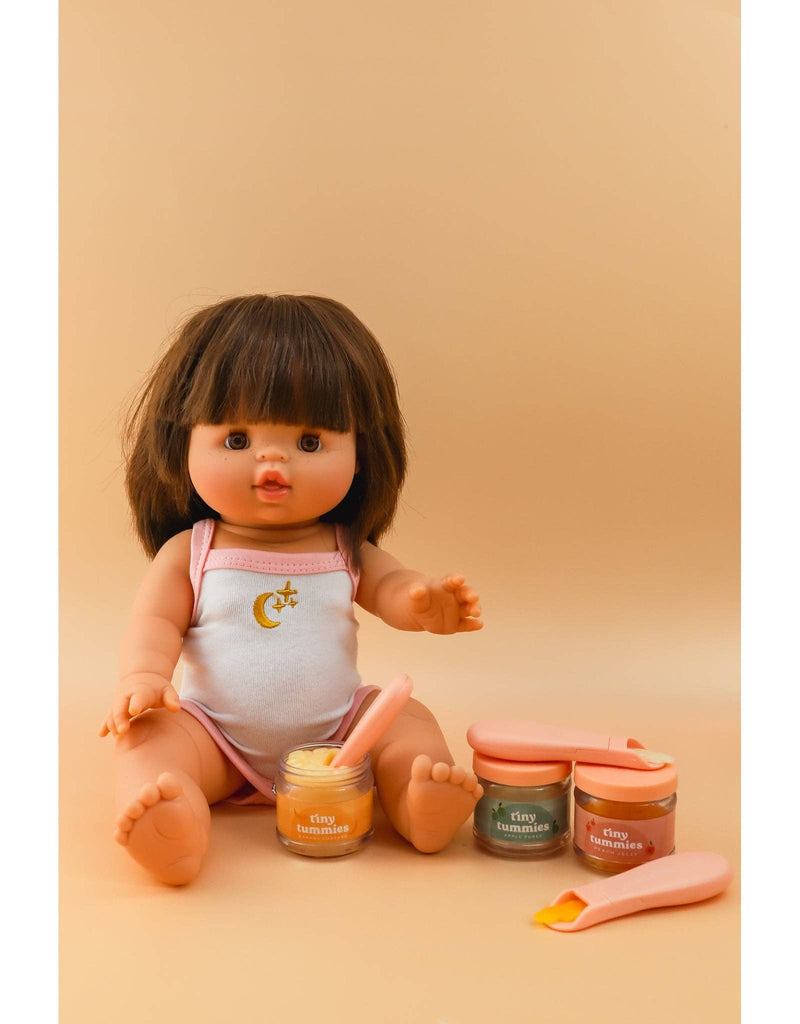 A doll with brown hair sits on a beige background, dressed in a white and pink outfit adorned with a crescent moon and star. In front are three jars of Tiny Tummies Banana Jelly food - Tiny Harlow, featuring pastel lids, each accompanied by a magic spoon.