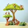 Wooden toy animals are displayed in a jungle setting with two trees. A Bumbu Wooden Blue Macaw Parrot sits on one tree while a monkey perches on the other. Two small rodents face each other on a branch. On the ground, two tigers and a tiger cub rest near green foliage. This Montessori & Waldorf inspired set is crafted from eco-friendly materials.