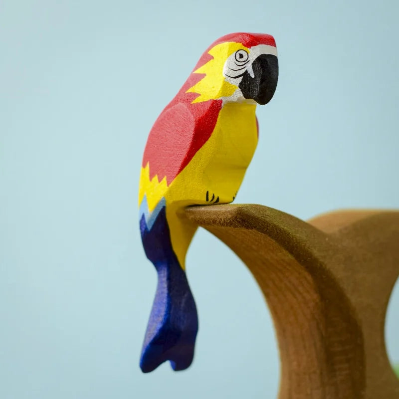 A vibrant Bumbu Wooden Amazonian Parrot figurine perched on a curved wooden stand. The parrot, adorned in bright red, yellow, blue, and black with intricate feather and facial details, is crafted from eco-friendly materials. It serves as a delightful Montessori educational toy against the pale blue background.