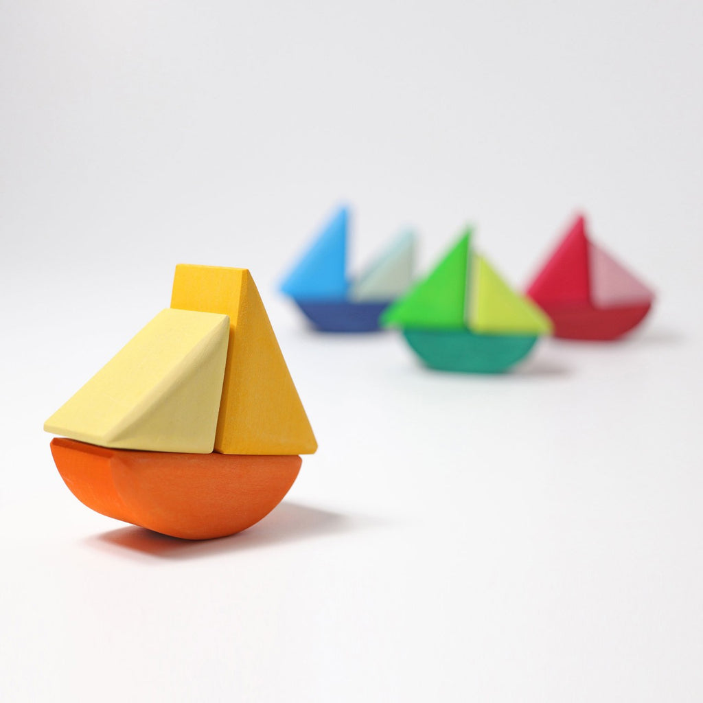 Four small and colorful Grimm's Rolling Boats are arranged in an angled line on a white surface. The closest boat is orange with yellow accents, followed by boats in blue, green, and red, each featuring contrasting colored sails. Perfect for small children, these non-toxic toys are safe and fun for imaginative play against a plain white wall.