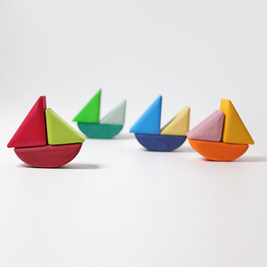 Four Grimm's Rolling Boats, each brightly painted in red, green, blue, and orange, are arranged on a white background. These colorful wooden toy sailboats are made of non-toxic materials and feature simple geometric shapes. Each boat has a half-circle base and a triangle sail—perfectly safe for small children to enjoy.