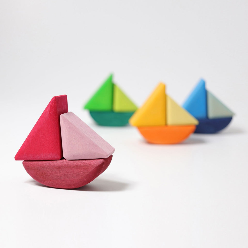 Four Grimm's Rolling Boats with minimalist designs are arranged in a row on a white background. Suitable for small children, the nearest boat features a red and pink color scheme, followed by green, yellow, and blue boats. Each boat is crafted from non-toxic materials and consists of simple geometric shapes that stack and combine easily.