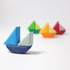 Four Grimm's Rolling Boats on a white surface. Closest to the forefront is a blue boat; farther back are boats in green, yellow, and red. All boats are simple, non-toxic, minimalist, and brightly colored, creating a playful scene perfect for small children to stack and combine.