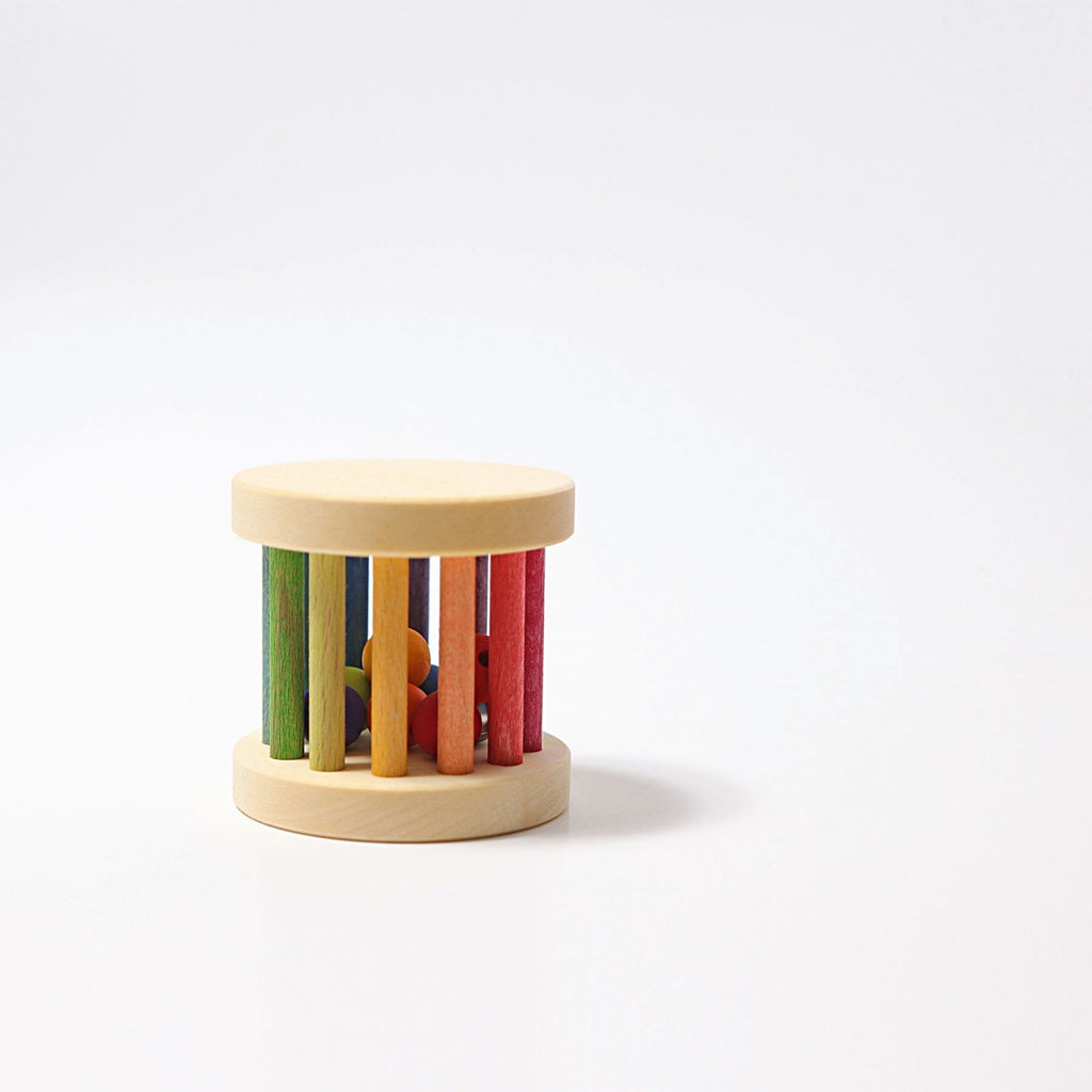 Mini Rolling Wheel featuring a cylindrical shape with vertical rainbow-colored bars and two circular wooden ends. Inside the Mini Rolling Wheel are small, brightly colored shapes designed to make sounds when shaken. The background is plain white.