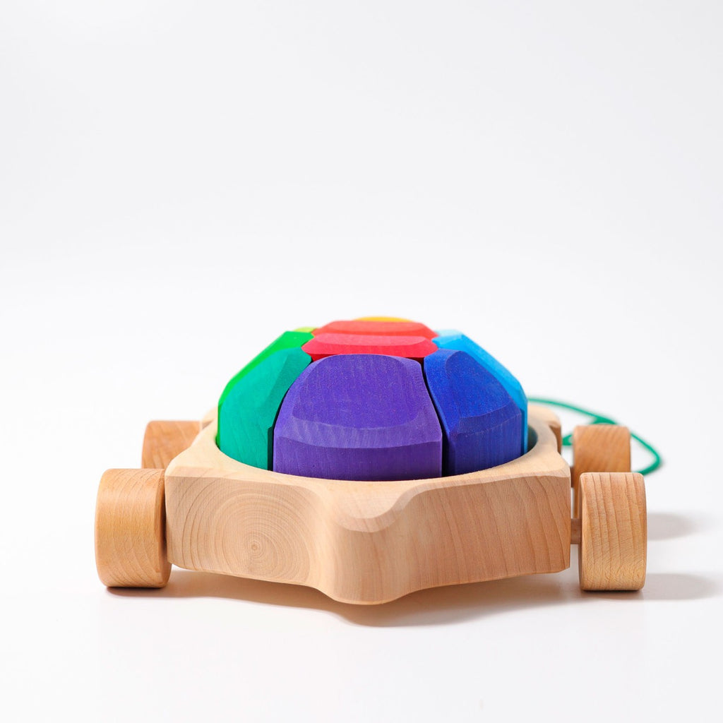 A Grimm's Rainbow Turtle Pull-Toy and Building Blocks with wheels featuring a dome made of colorful, segmented shapes in blue, purple, green, and red on top. The dome is mounted on a natural wood base with four wheels and a green string attached for pulling. Made from non-toxic materials to enhance fine motor skills, the toy is set against a plain white background.