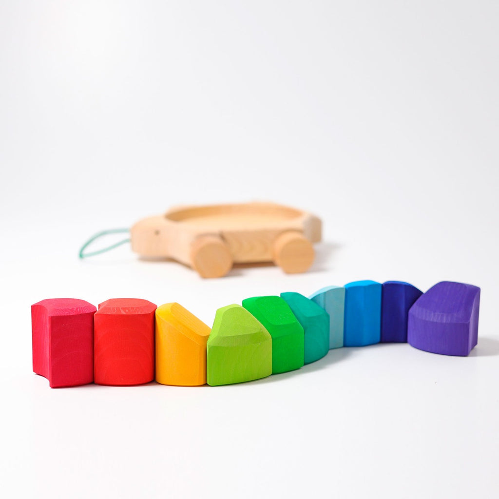 A set of Grimm's Rainbow Turtle Pull-Toy and Building Blocks arranged in a line, featuring hues of red, orange, yellow, green, blue, and purple. In the background, there is a light-colored wooden pull toy with wheels and a light blue string attached. Both items are crafted from non-toxic materials to help develop fine motor skills.