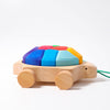 The Grimm's Rainbow Turtle Pull-Toy and Building Blocks is shaped like a turtle with a shell made of colorful, removable blocks, crafted from non-toxic materials. The toy features wheels and a green string attached to the front for pulling. The vibrant shell blocks in shades of blue, red, yellow, and green help develop fine motor skills.