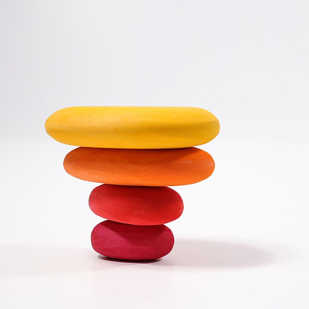 Four smooth, organically shaped stones are stacked atop each other against a plain white background. From bottom to top, the stones are colored red, deep orange, light orange, and yellow with non-toxic color stain, creating a visually striking gradient effect. This arrangement is achieved using the Grimm's Fire Pebbles Stacker.