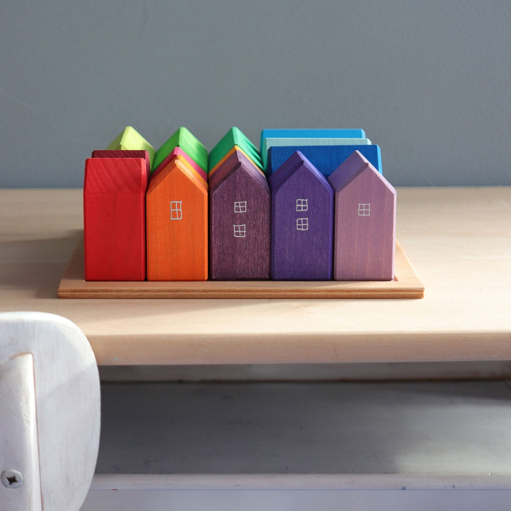 A delightful display of Grimm's Houses, hand-painted in vibrant colors and crafted from non-toxic materials, is neatly arranged on a light wood shelf. Each wooden block features a distinct hue—red, orange, green, blue, and purple—and charming window patterns that evoke the feel of picturesque stops along an imaginative wooden train route.