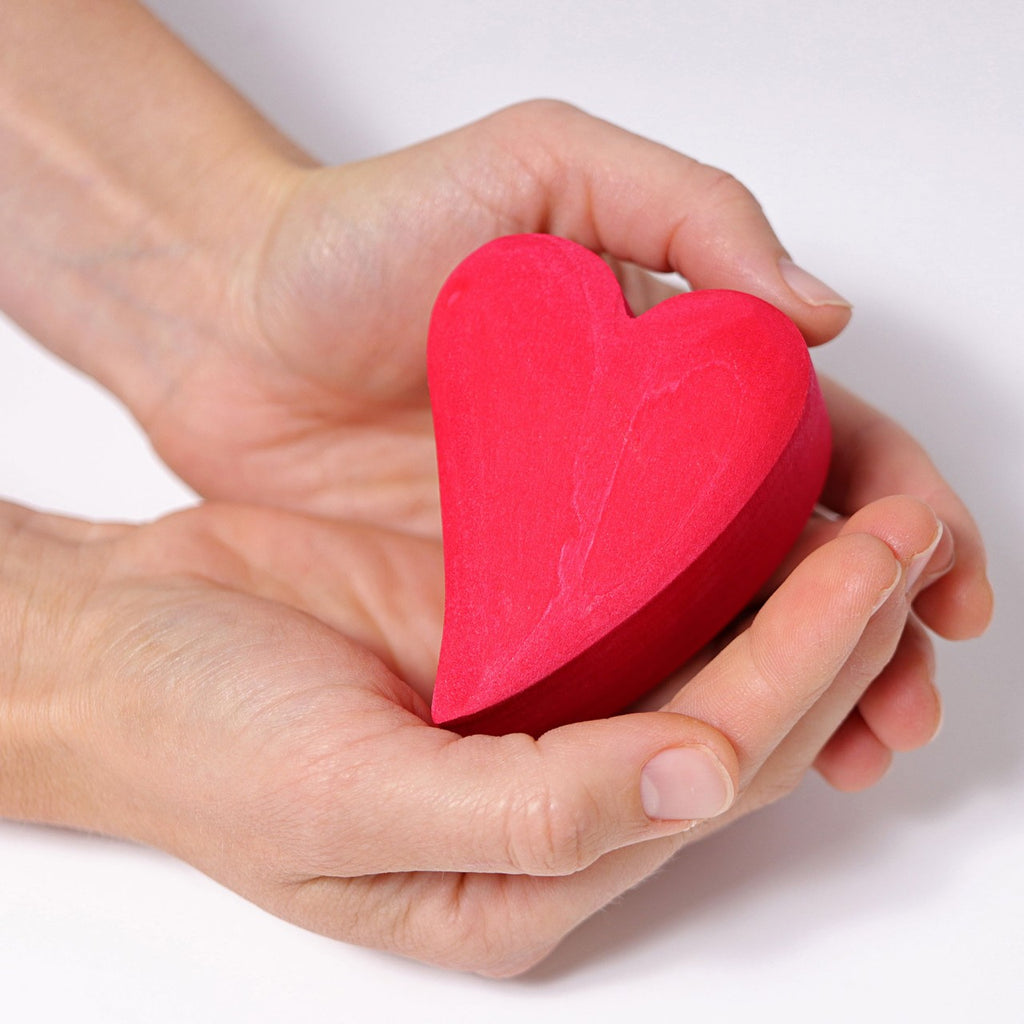 Two hands are gently holding a Grimm's Red Hearts Building Blocks, reminiscent of a crafted desk-toy. The background is plain and white, emphasizing the vibrant color and shape of the heart. The hands are positioned cupped together, conveying a sense of care and protection.