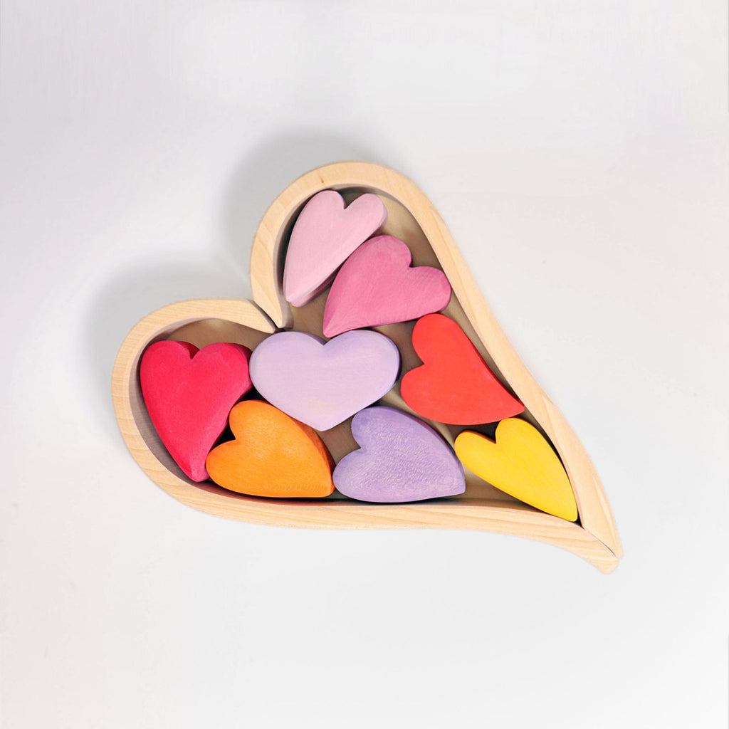 A wooden heart-shaped frame contains nine smaller, variously colored hearts. These hearts, in shades of pink, red, orange, purple, and yellow, are arranged in an aesthetically pleasing pattern against a plain white background. Perfect as a desk toy or to enhance fine motor skills with its colorful building blocks. Introducing Grimm's Red Hearts Building Blocks.