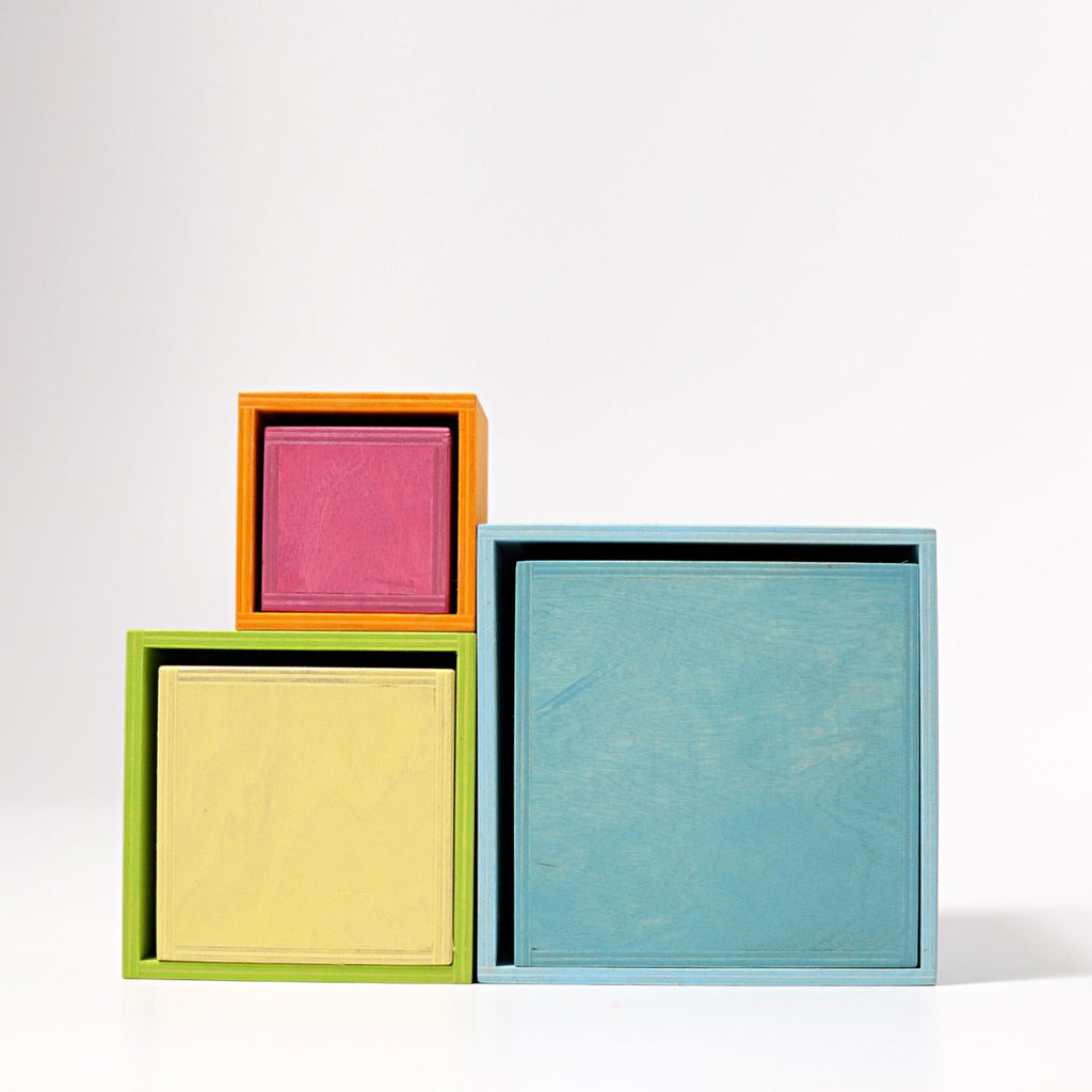 Four nested stacking boxes of different sizes and colors are stacked. From left to right: a small orange and pink box, a medium green and yellow box, and a large blue box. Made from non-toxic wood, this delightful children's toy sets against a white background. The product is Grimm's Large Pastel Set of Boxes.