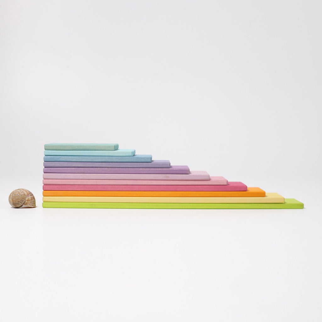 A neatly arranged sequence of colorful, rectangular wooden blocks of varying sizes is displayed against a white background. These Grimm's Pastel Building Boards Set, perfect for open-ended play, are stacked from largest to smallest, left to right, featuring various shades of green, blue, purple, pink, orange, and yellow.