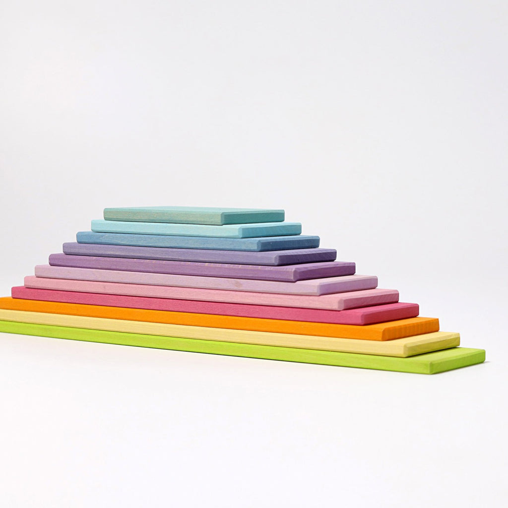 A neatly stacked pyramid of Grimm's Pastel Building Boards Set in various vibrant colors. The blocks are arranged in a gradient from bottom to top, starting with bright green and progressing through yellow, orange, red, pink, purple, blue, and light blue—perfect for open ended play and skill games.